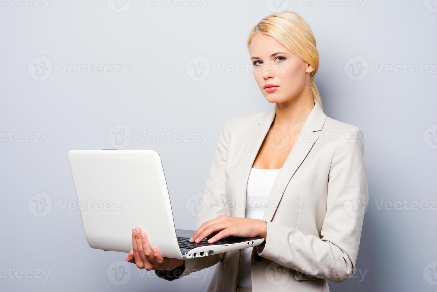 Supporting your business. Confident young businesswoman holding laptop while standing against grey background photo