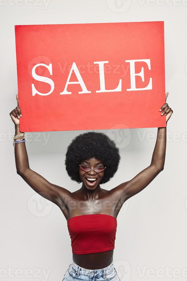 Happy young African woman holding sale banner and smiling while standing against gray background photo