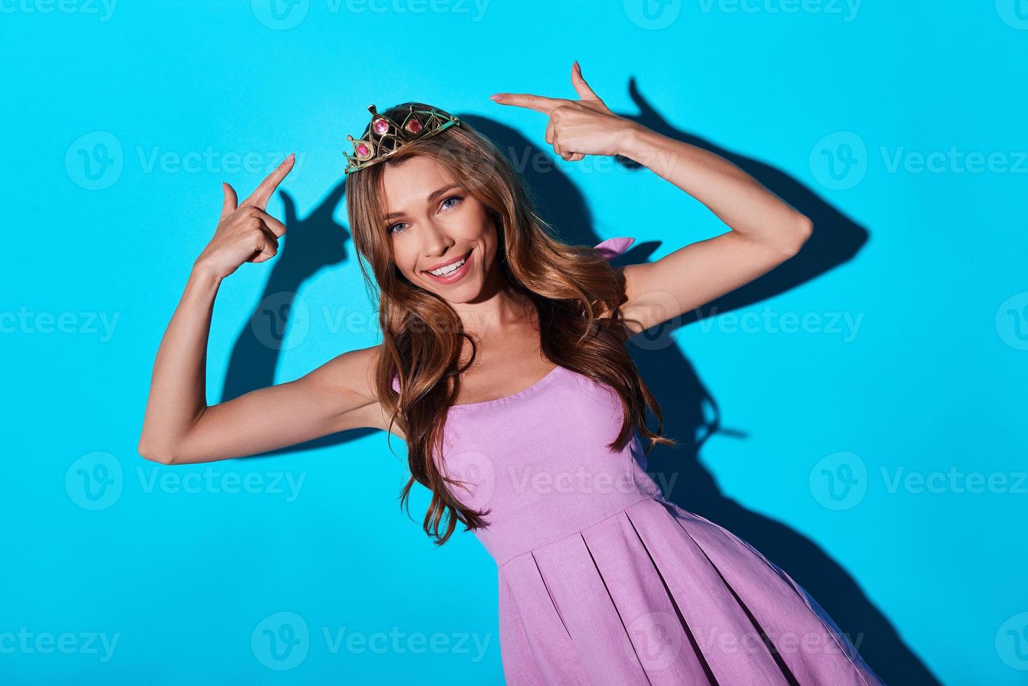 glamour. atractiva mujer joven señalando su tiara y sonriendo mientras está de pie contra el fondo azul foto