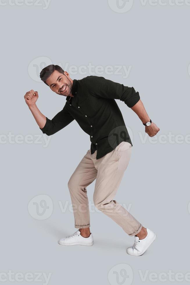 Going crazy. Full length of playful young man dancing and smiling while standing against grey background photo