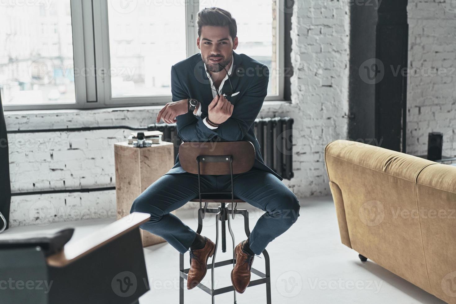 Charming man. Handsome young man in full suit looking at camera and smiling while sitting on the stool photo