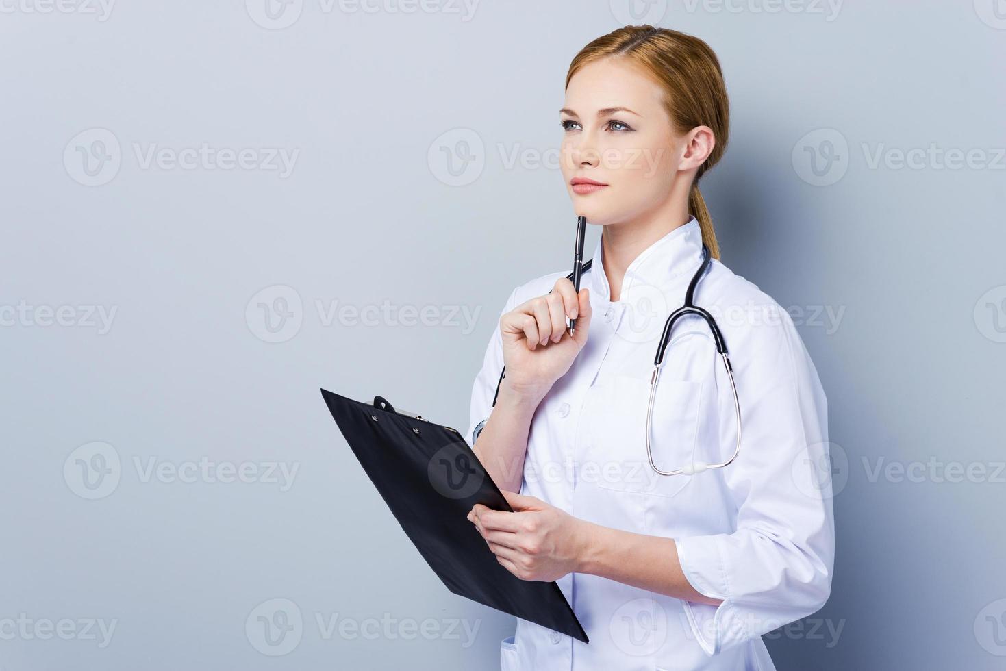 Leading an important medical documentation. Confident female doctor in white uniform holding clipboard while standing against grey background photo