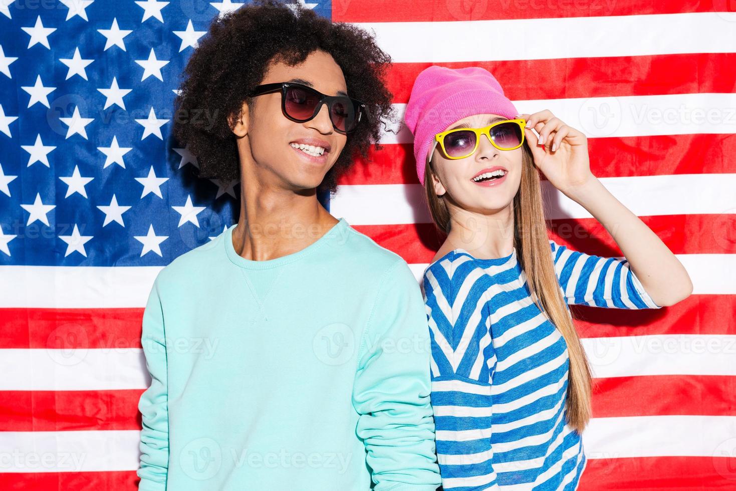 sueños americanos funky pareja joven con gafas de sol y sonriendo mientras está de pie contra la bandera americana foto