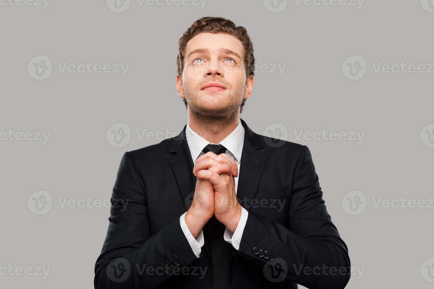 hombre de negocios rezando. un joven pensativo vestido con ropa formal con las manos entrelazadas y los dedos cruzados mientras se enfrenta a un fondo gris foto