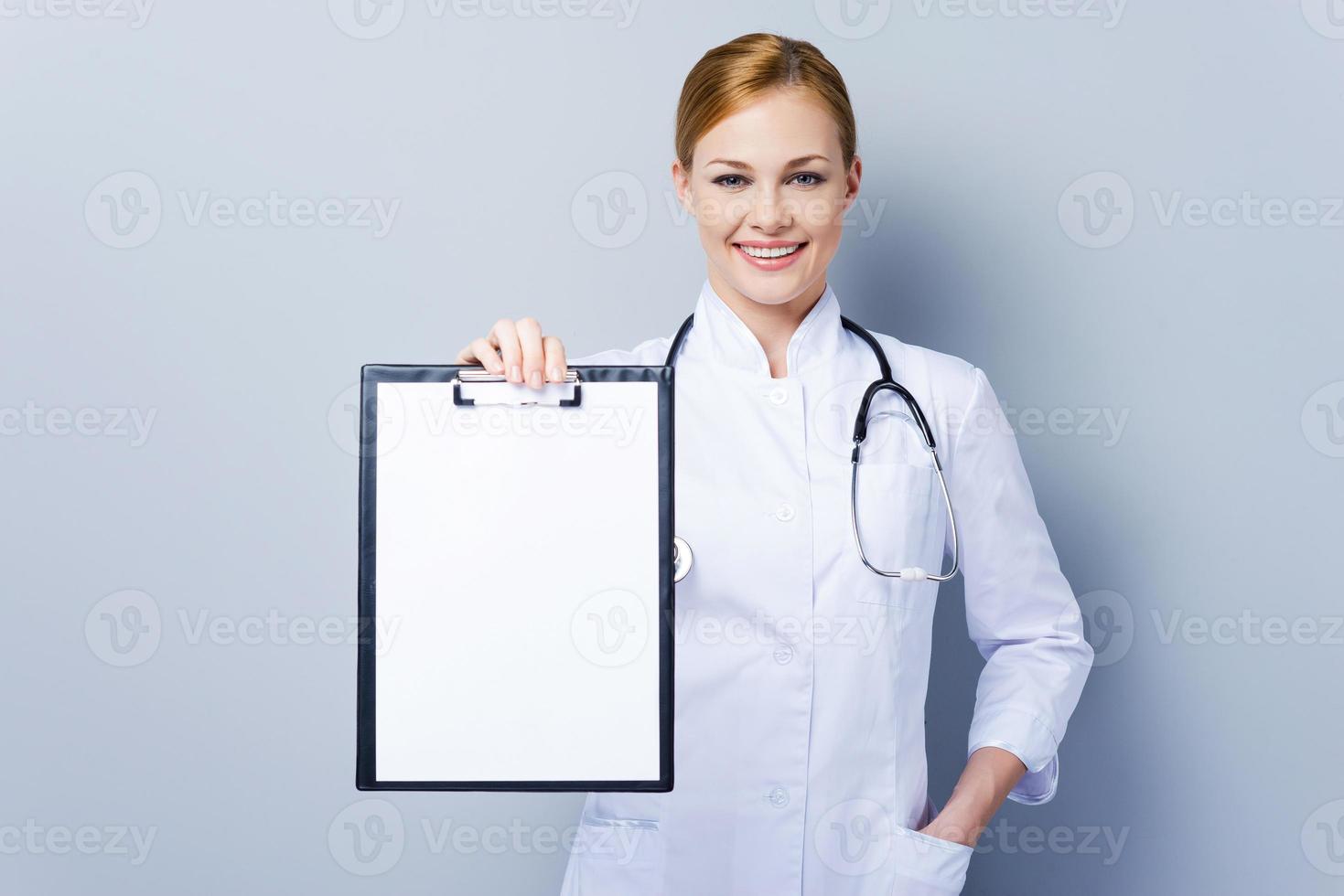 Copy space on her clipboard. Confident female doctor in white uniform holding clipboard and looking at camera while standing against grey background photo