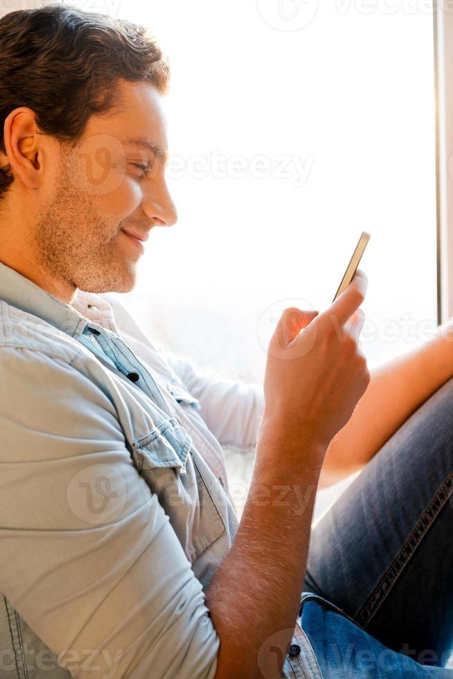 Good news from friend. Side view of handsome young man sitting at the window sill and holding mobile phone photo