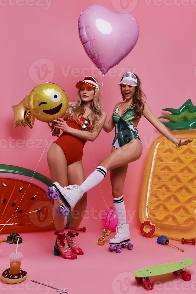 Girls having fun.  Full length of two playful young women in swimwear bonding and smiling while standing against pink background photo