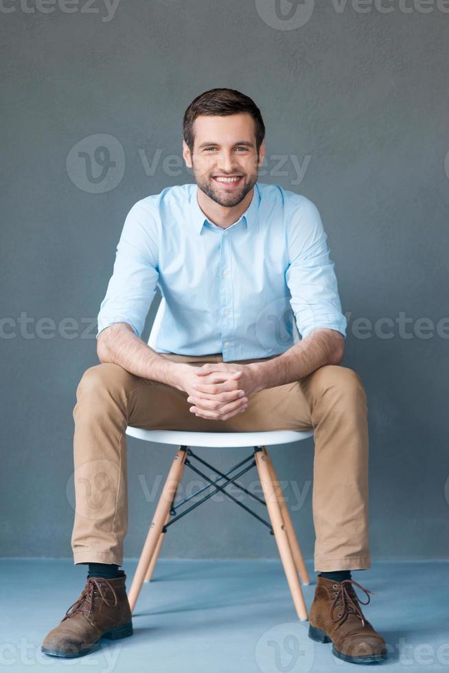 Positive thinker. Full length of handsome young man looking at camera and smiling while sitting against grey background photo