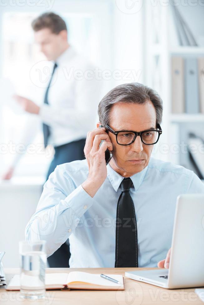 Business time. Confident businessman in formalwear working on laptop and talking on the mobile phone while sitting at desk in the office photo