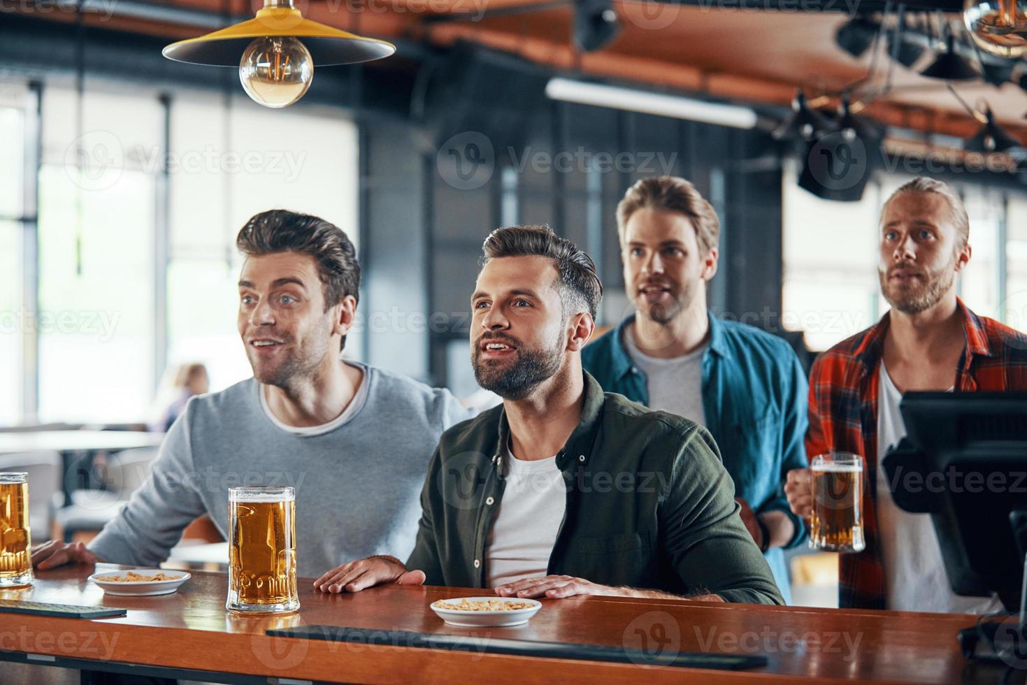 Cheering young men in casual clothing watching sport game and enjoying beer while sitting in the pub photo