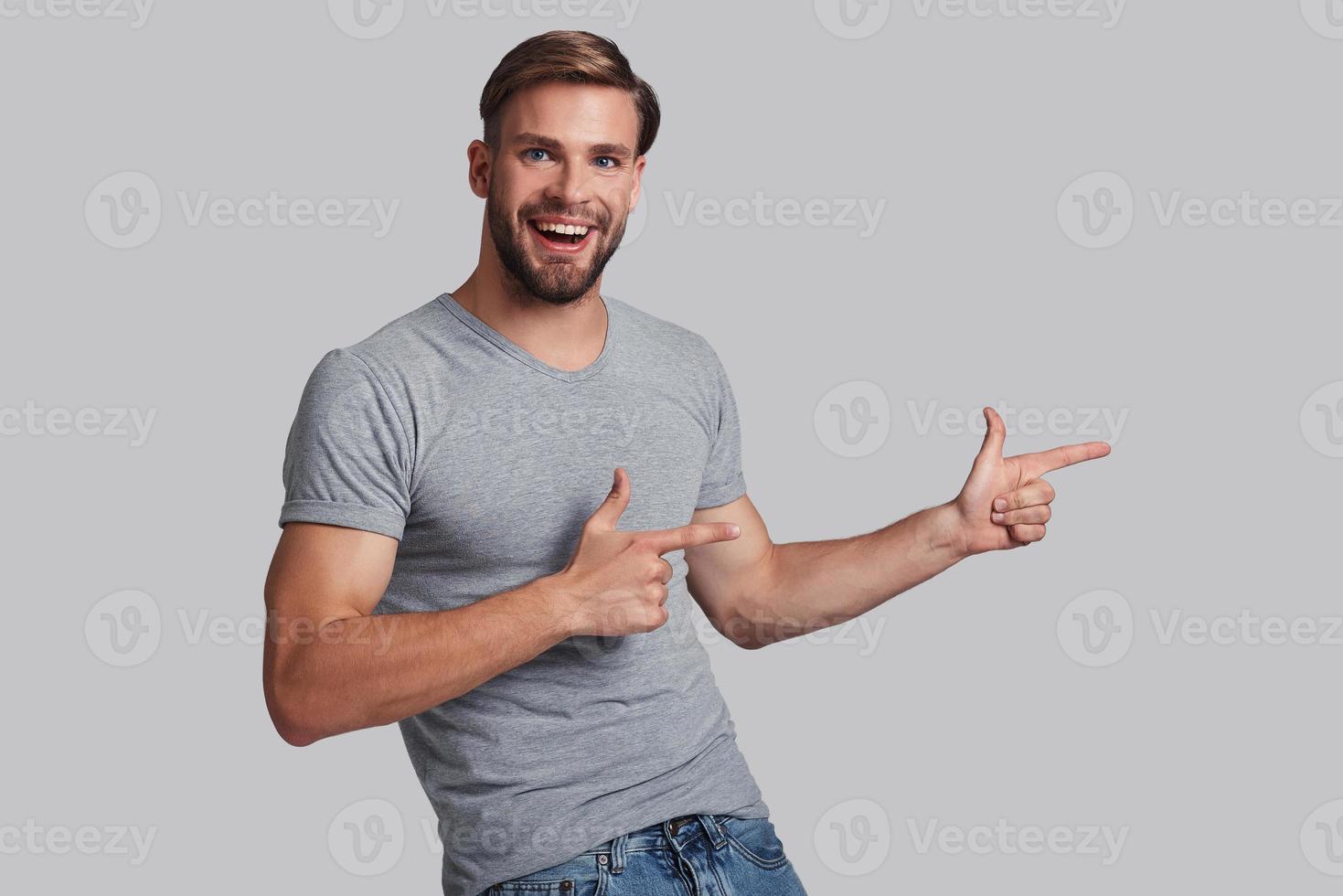 Take a look over here. Charming young man pointing copy space and smiling while standing against grey background photo