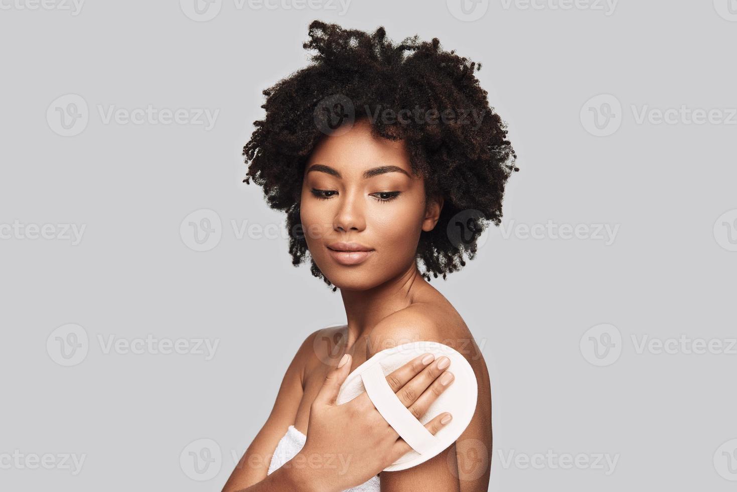 piel suave y limpia. atractiva joven africana aplicando esponja de limpieza y sonriendo mientras está de pie contra el fondo gris foto