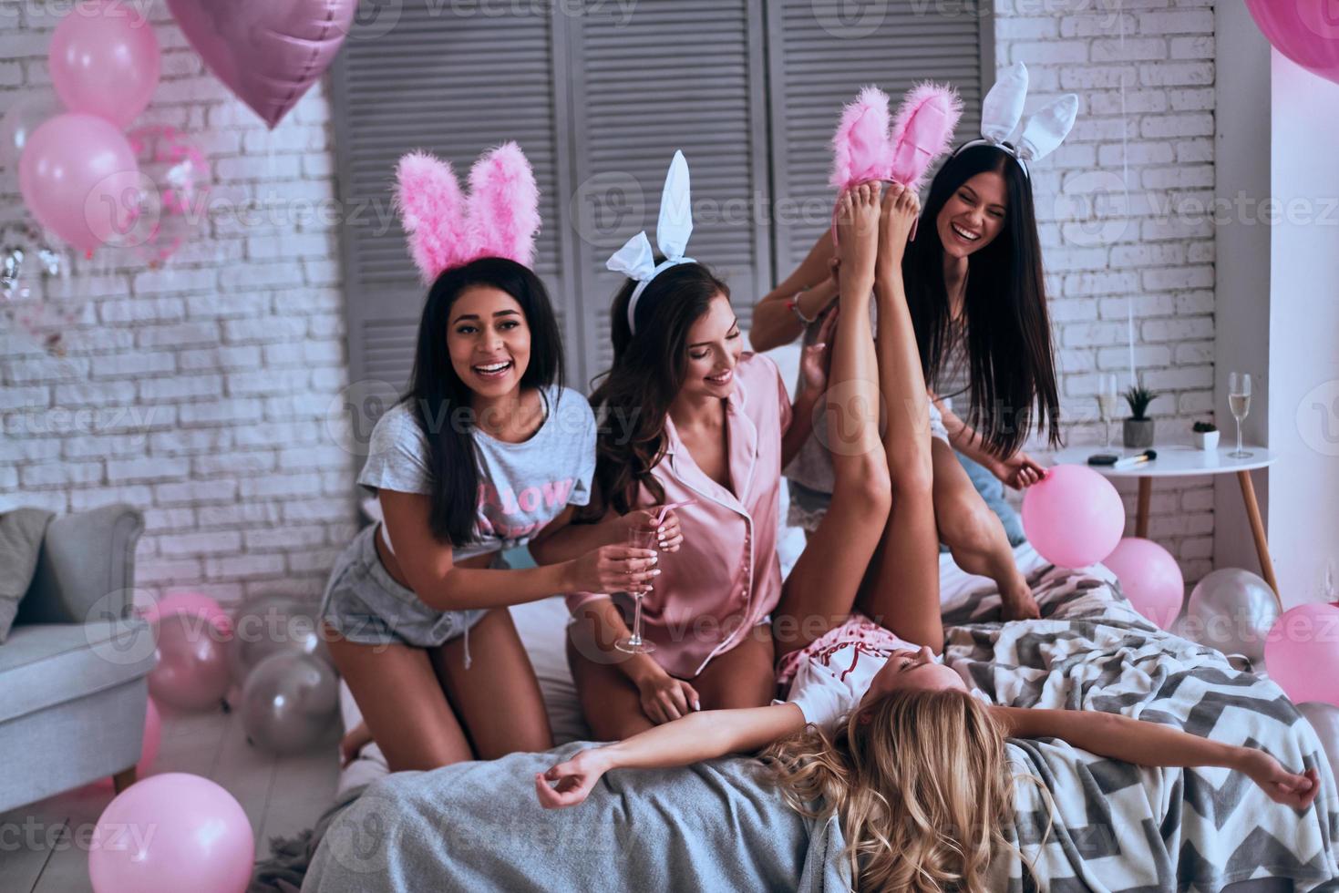 haciendo lo que quieran. mujeres jóvenes juguetonas tratando de ponerse orejas de conejo en los pies de sus novias y sonriendo mientras se sientan en la cama foto