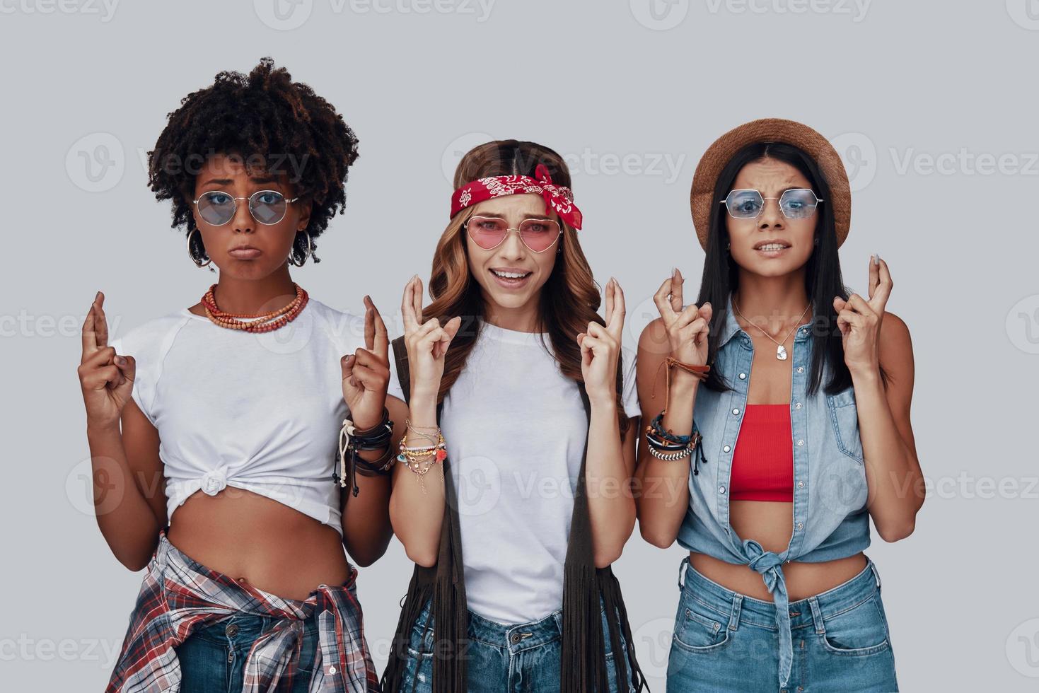 Three attractive young women keeping fingers crossed and looking at camera while standing against grey background photo