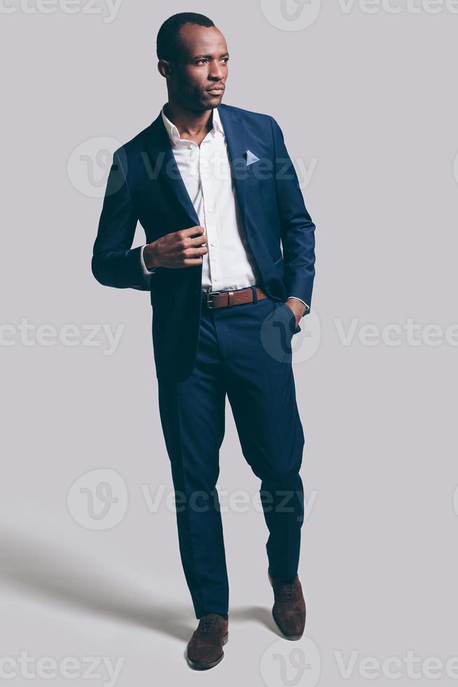 Confident and stylish. Full length of handsome young African man in full suit adjsuting his jacket and holding one hand in pocket while standing against grey background photo