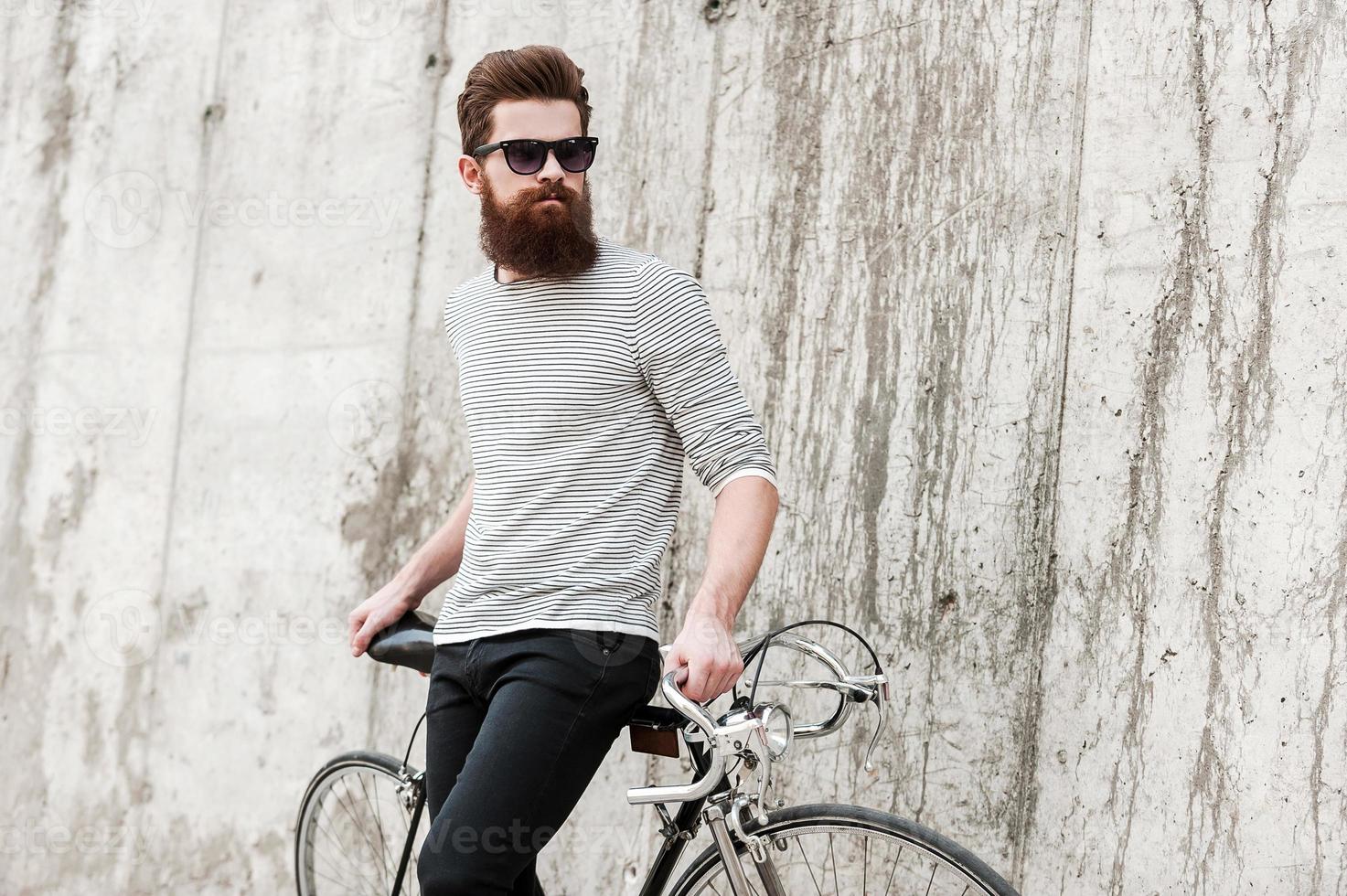 Street style. Pensive young bearded man leaning at his bicycle and looking away while standing against the concrete wall photo