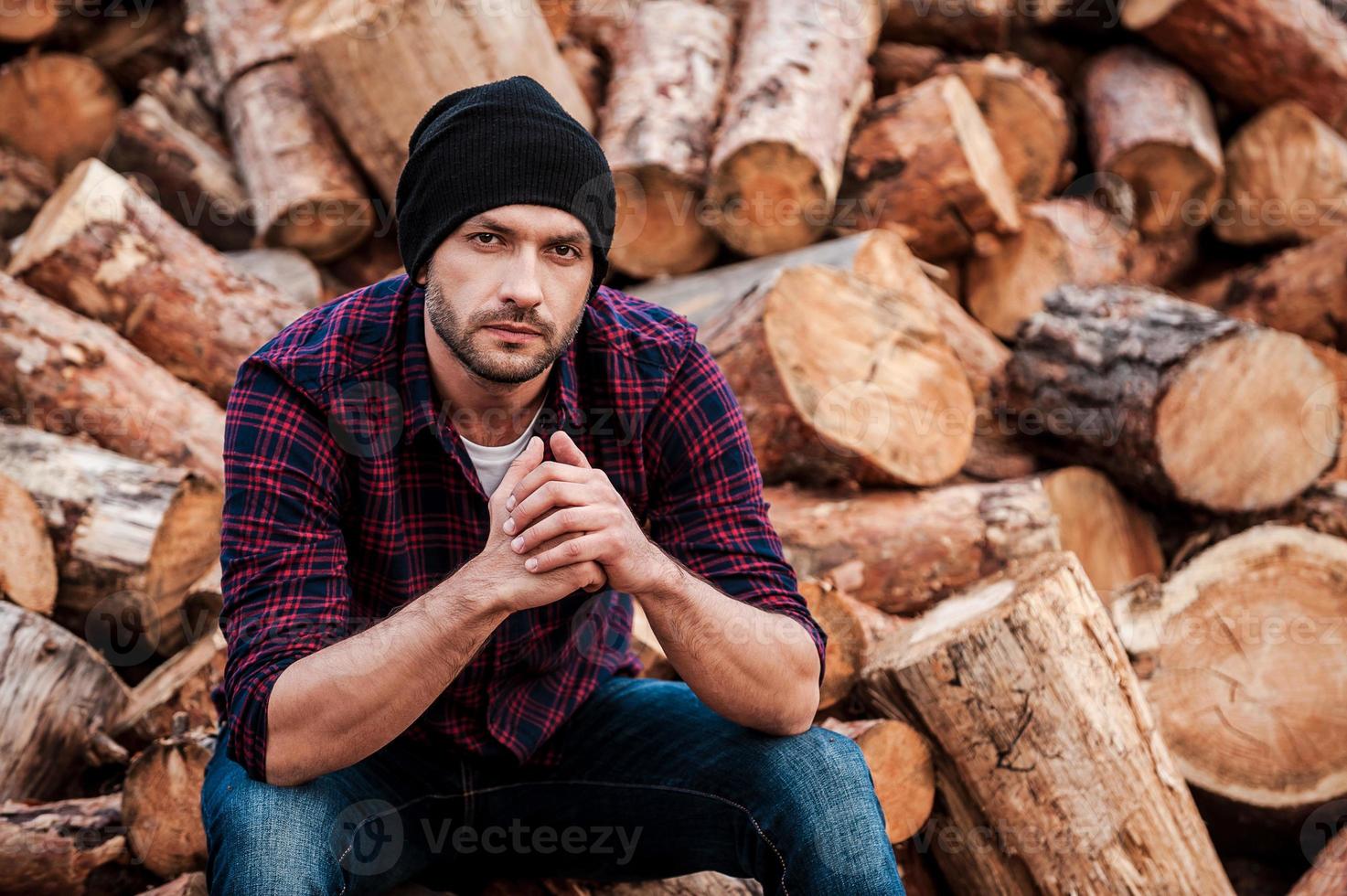 Day dreaming on the logs. Handsome young forester keeping hands clasped and looking at camera while sitting on logs photo