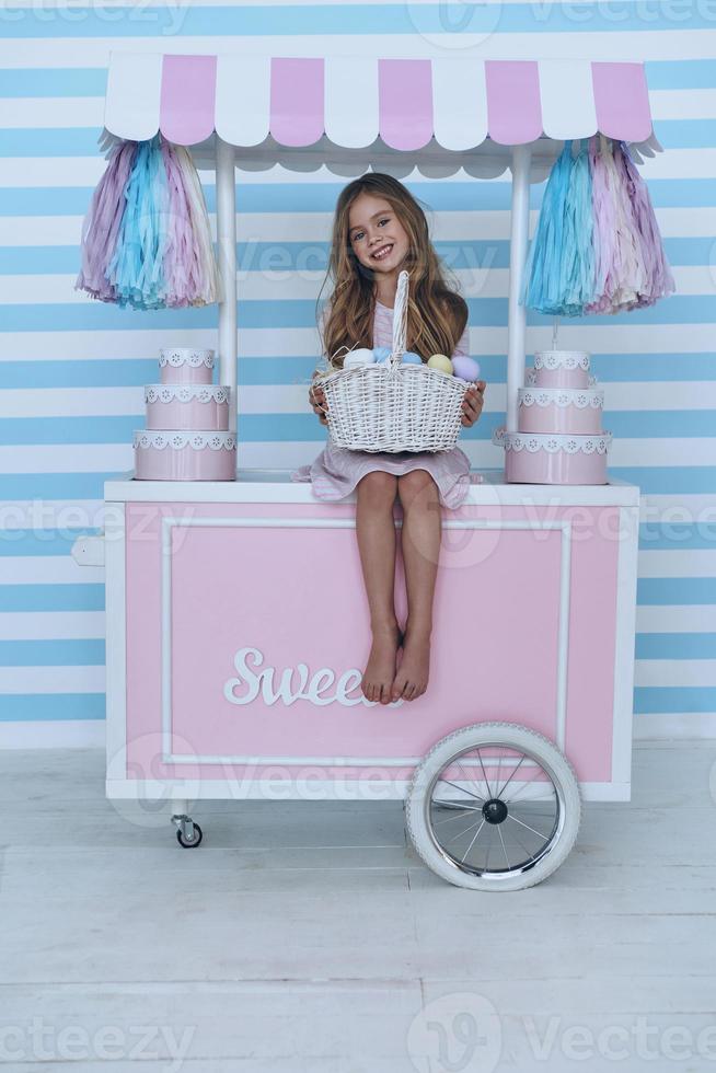 niño encantador linda niña sosteniendo una canasta de pascua y sonriendo mientras se sienta en la decoración del carrito de dulces foto