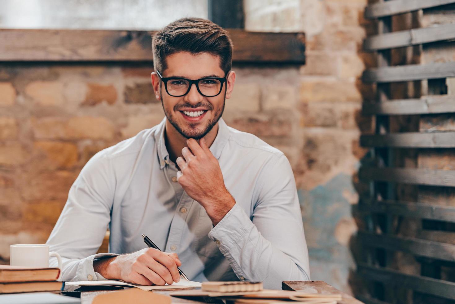 Working with pleasure. Handsome young man in glasses making some notes in his note pad and looking at camera with smile while sitting at his working place photo