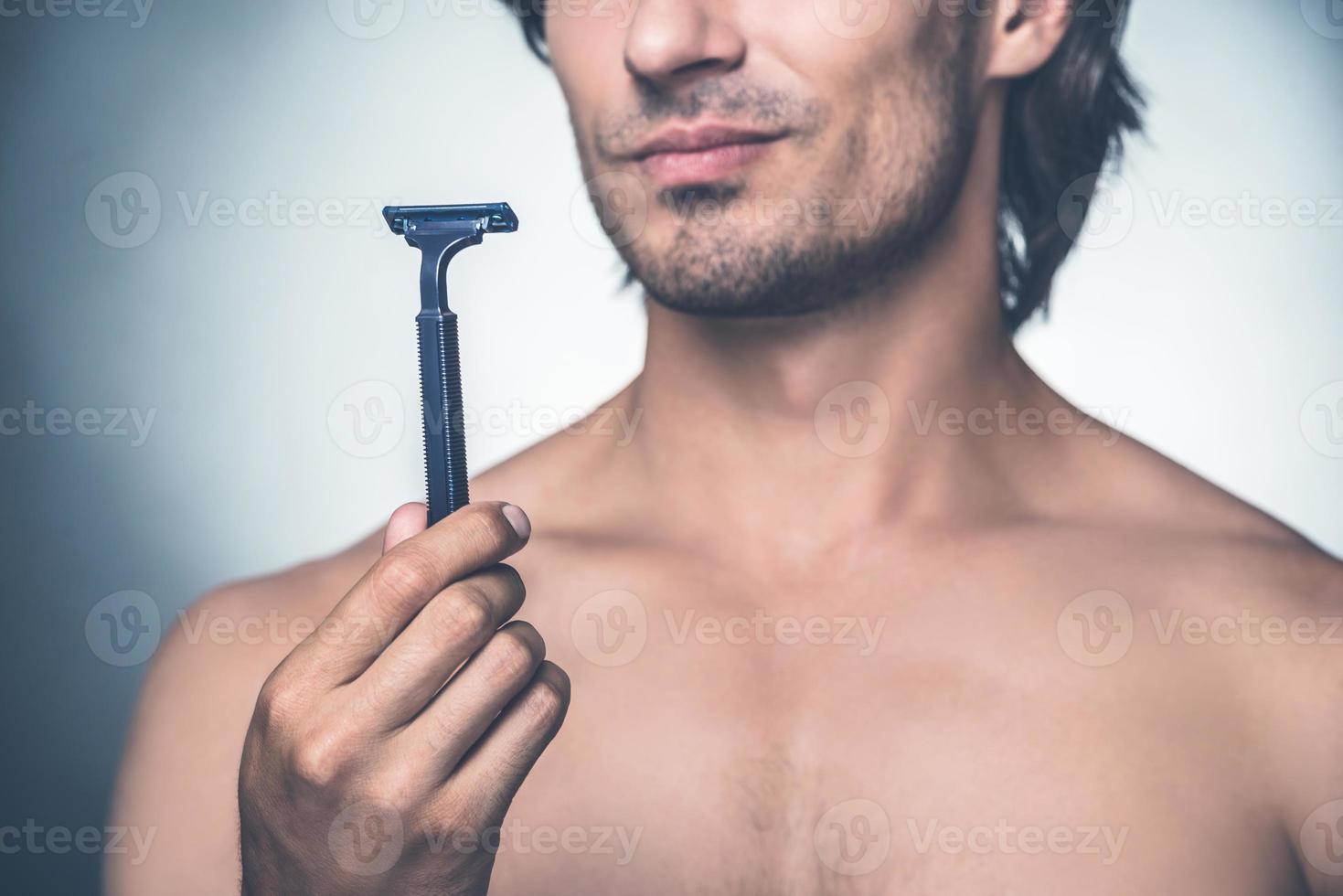 Time for new razor. Close-up young shirtless man holding razor and expressing negativity while standing against grey background photo