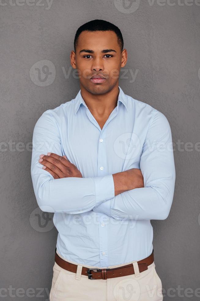 Confident and handsome. Confident young African man keeping arms crossed and looking at camera while standing against grey background photo