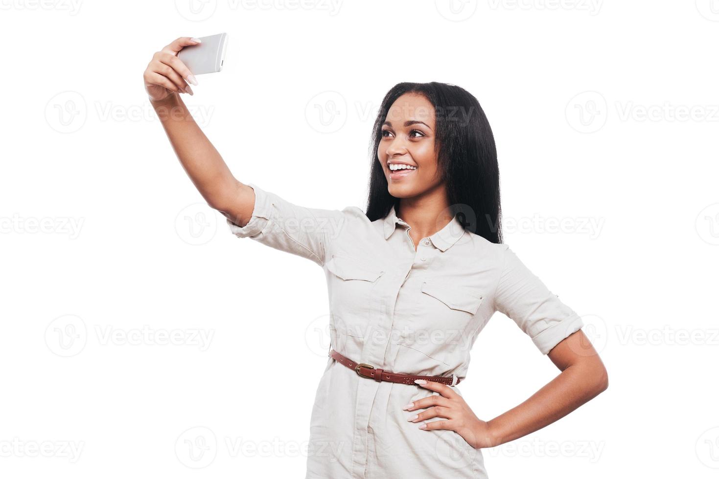 hora de hacerse un selfie. bella joven africana haciéndose selfie con su teléfono inteligente y sonriendo mientras se enfrenta a fondo blanco foto