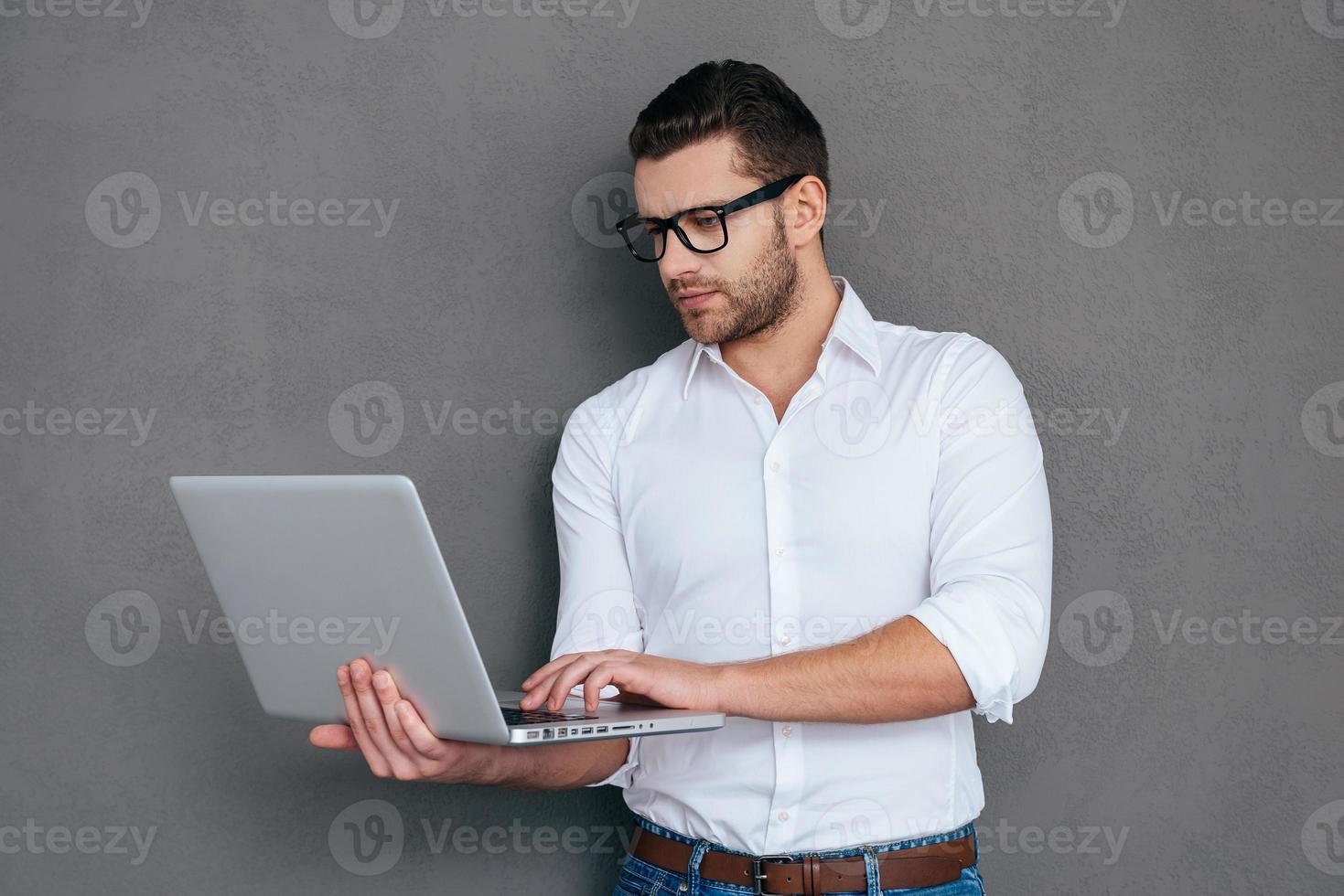 Man with laptop. Handsome young man working on laptop while standing against grey background photo