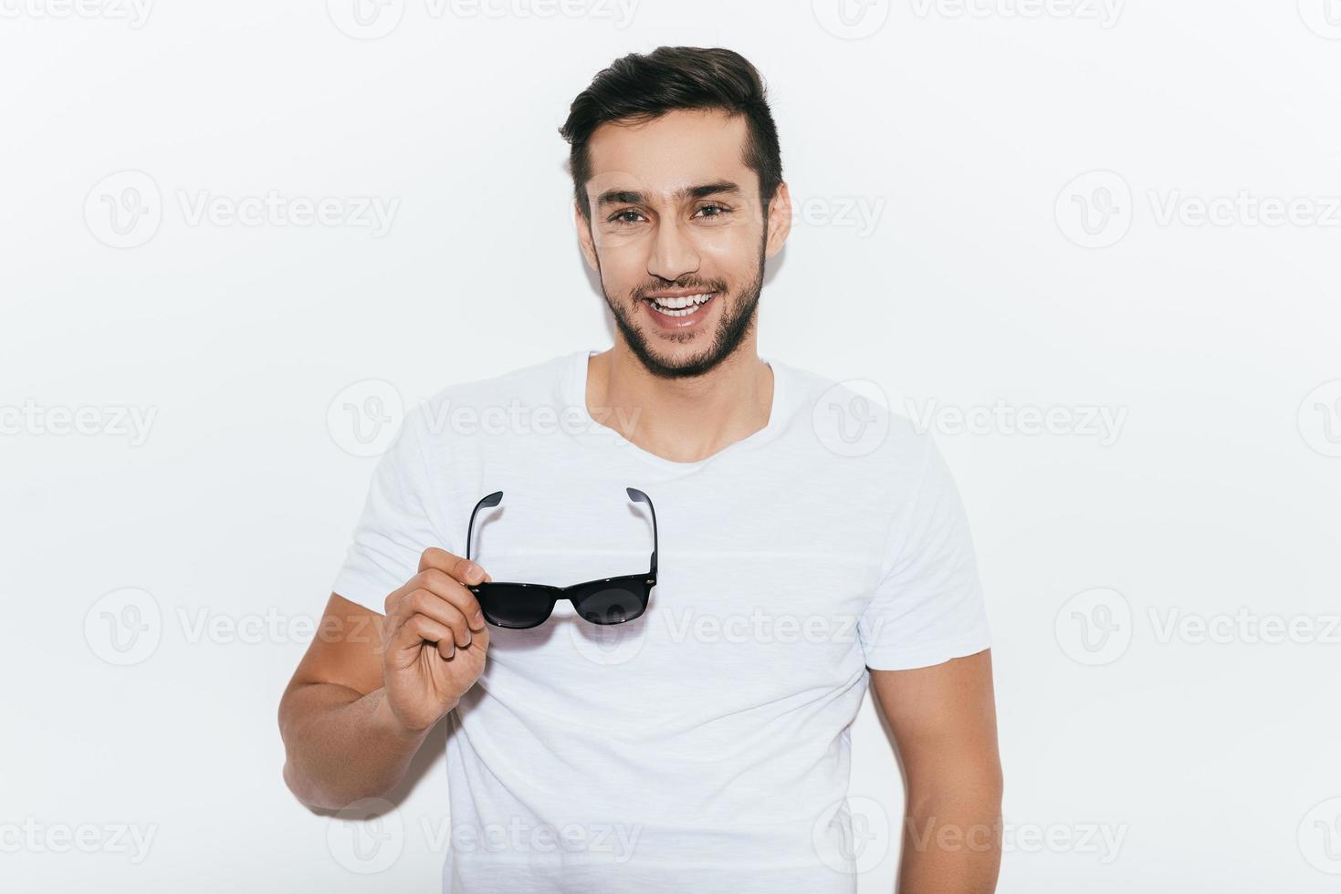 Charming heartbreaker. Handsome young Indian man carrying sunglasses and looking at camera with smile while standing against white background photo