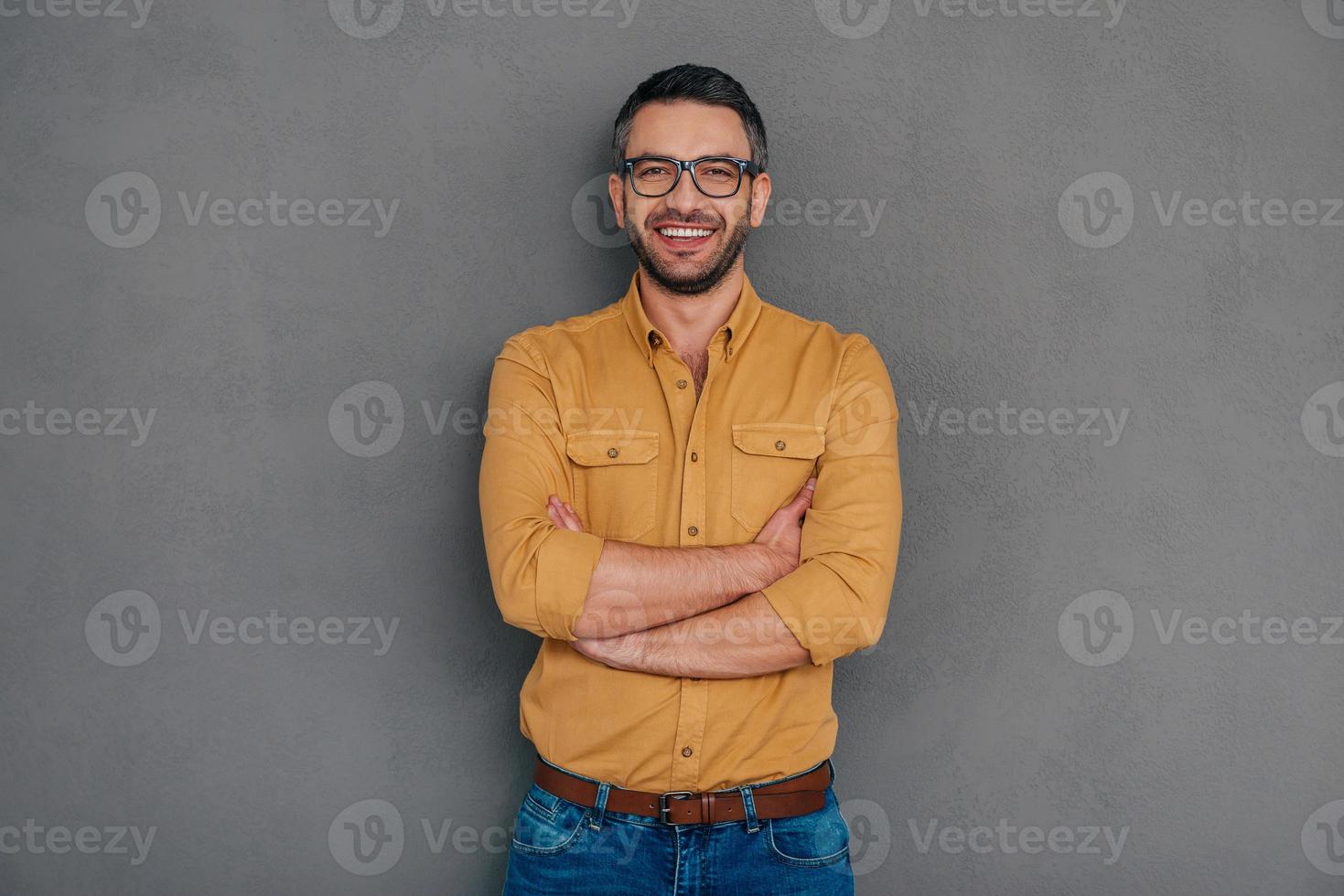 confiado y exitoso. hombre maduro seguro de sí mismo sosteniendo la mano en la barbilla y mirando a la cámara con una sonrisa mientras se enfrenta a un fondo gris foto