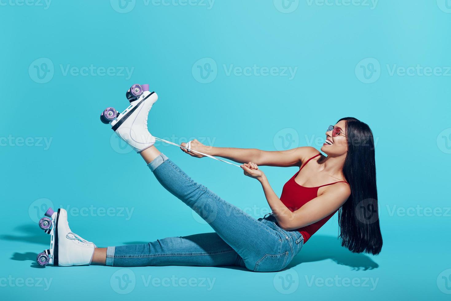 Attractive young woman tying roller skate and smiling while sitting against blue background photo