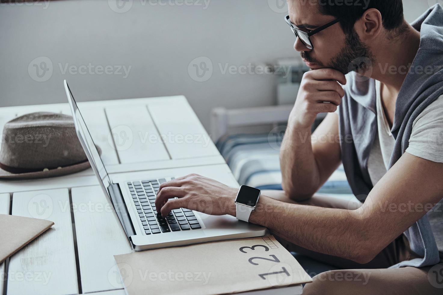 Feeling passionate about his project. Thoughtful young modern man using computer and keeping hand on chin while sitting on the sofa photo