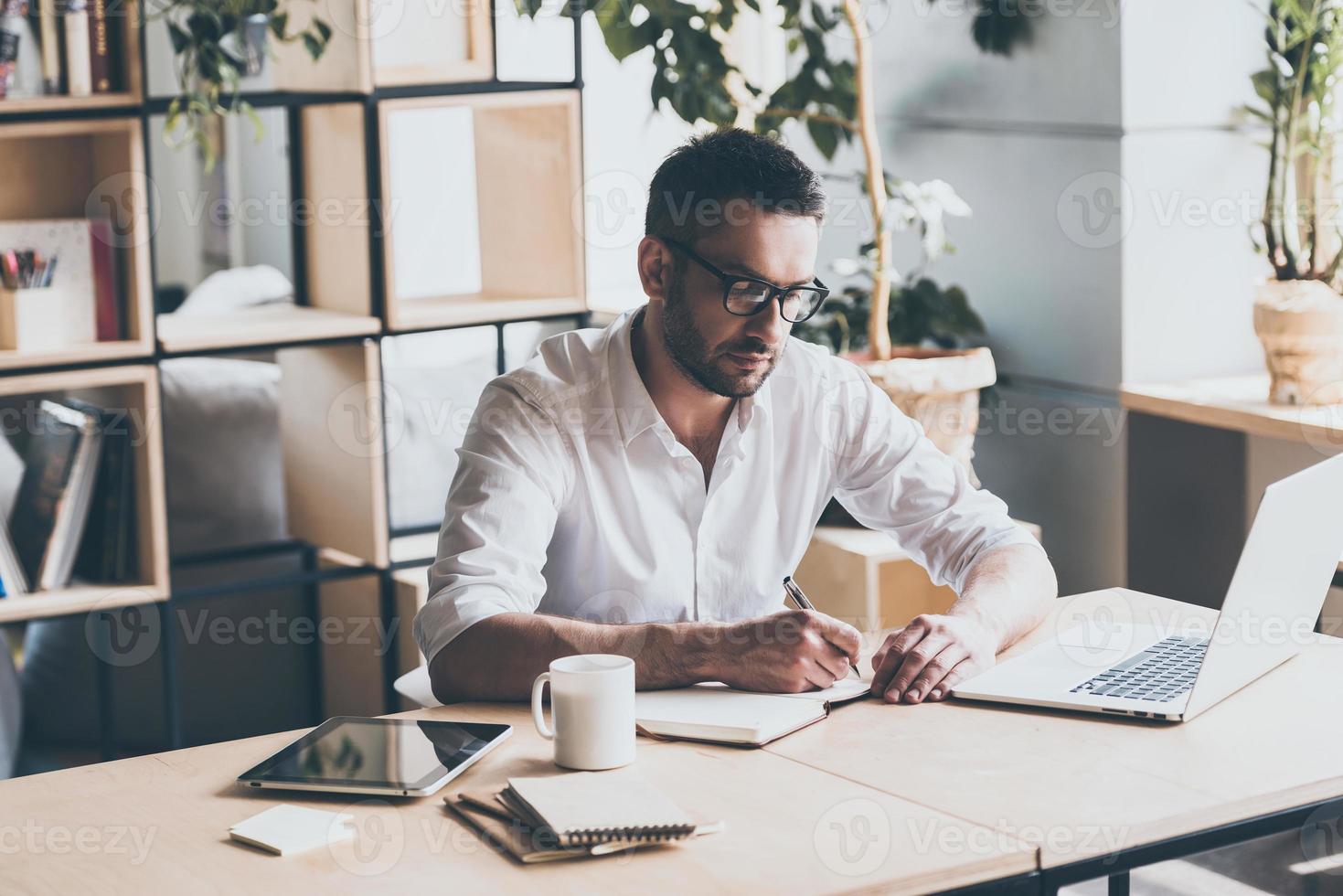 Making some notes. Concentrated mature man writing something in note pad while sitting at his working place in office photo