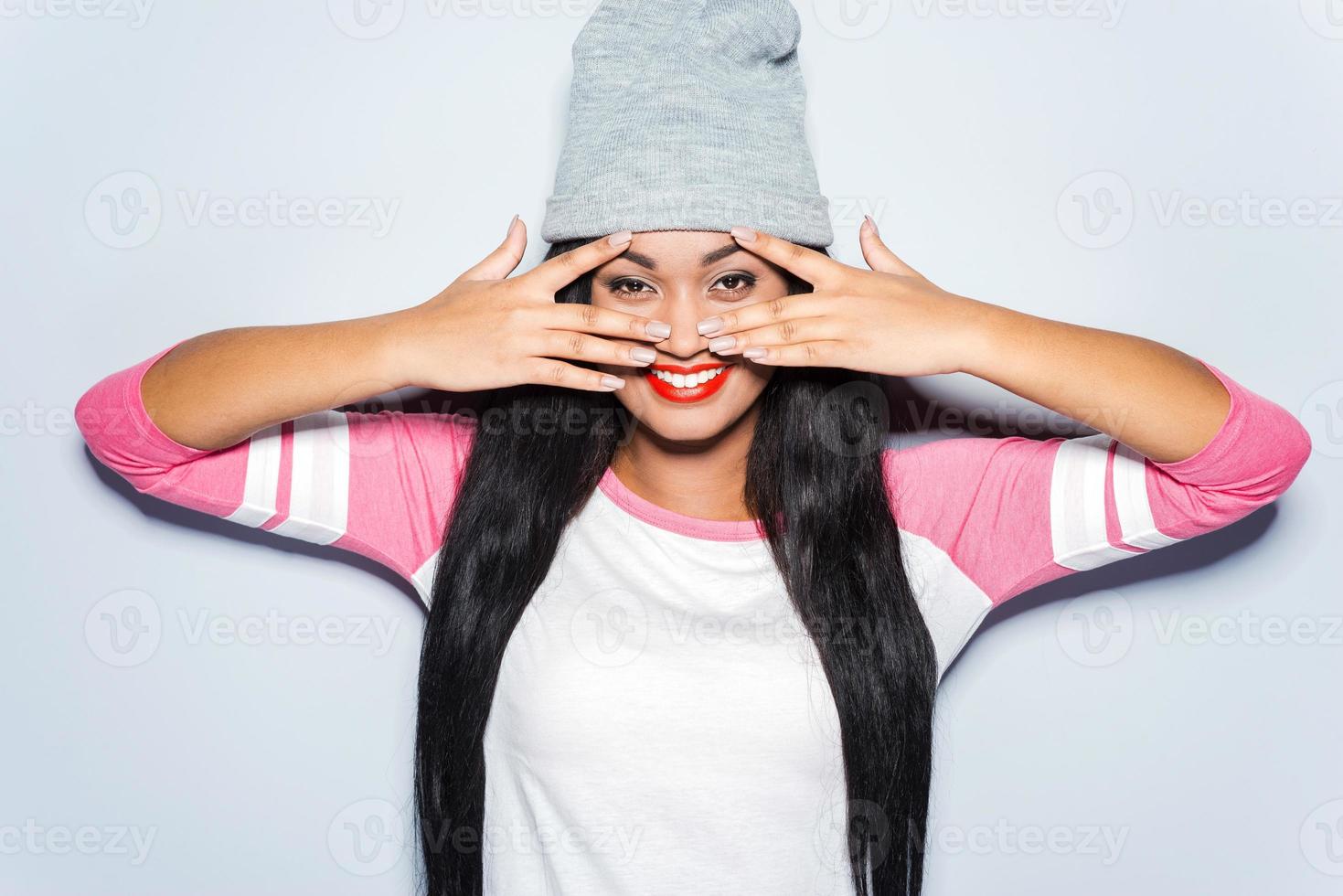 Just fun in my mind. Happy young African woman in funky clothes touching her face with fingers and smiling while standing against grey background photo