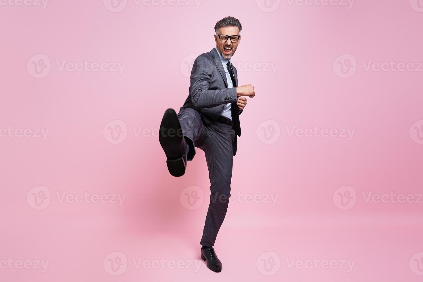 furioso hombre maduro con ropa formal lanzando una patada en la pierna mientras se enfrenta a un fondo rosa foto