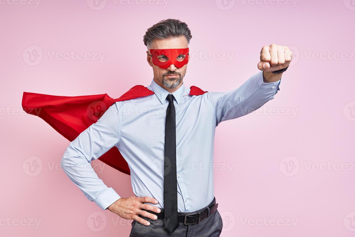 Confident mature man wearing superhero cape and keeping arm outstretched against pink background photo