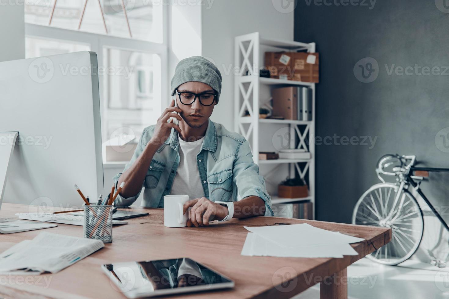 In touch with clients. Handsome young man in eyewear talking on smart phone while sitting at his working place in creative office photo