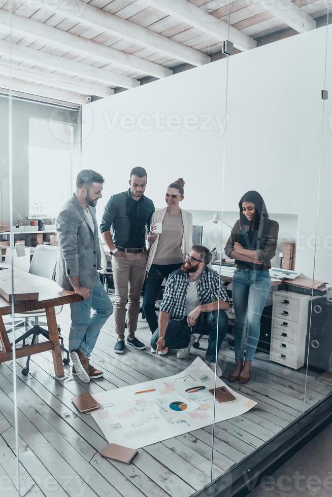 Successful business team. Group of young modern people in casual wear planning business strategy while standing behind the glass wall in the creative office and looking at large blueprint laying on the floor photo