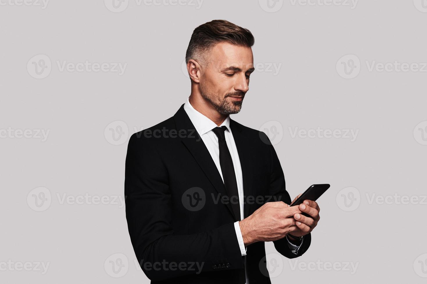 Always available. Good looking young man in full suit using his smart phone and smiling while standing against grey background photo