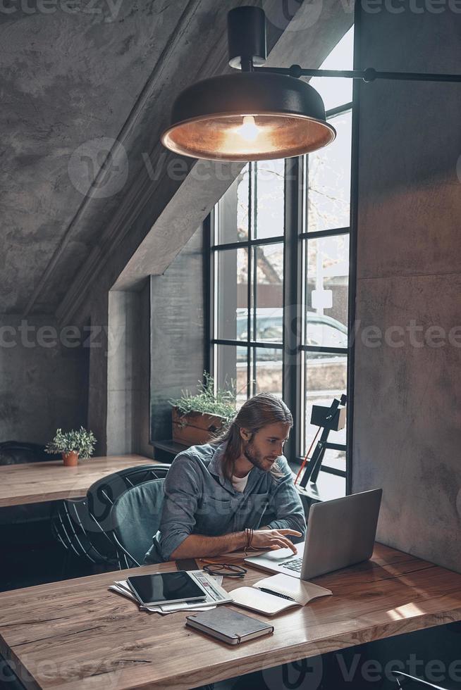 Feeling passionate about his project. Top view of thoughtful young man working using computer while sitting at the office desk near the window photo