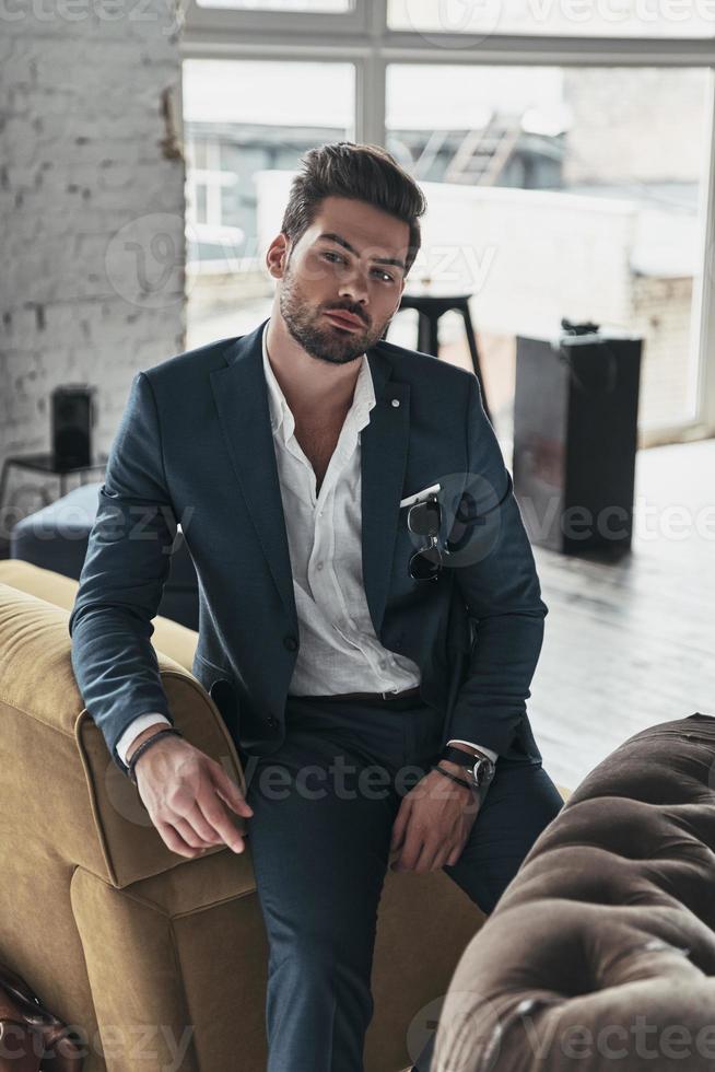 Elegance and masculinity. Thoughtful young man in full suit looking at camera while leaning on the sofa photo