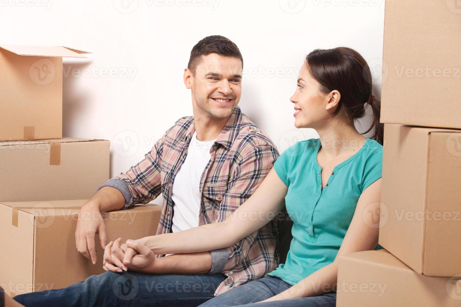 Just moved in a new apartment. Beautiful young loving couple sitting on the floor and holding hands while cardboard boxes laying around them photo