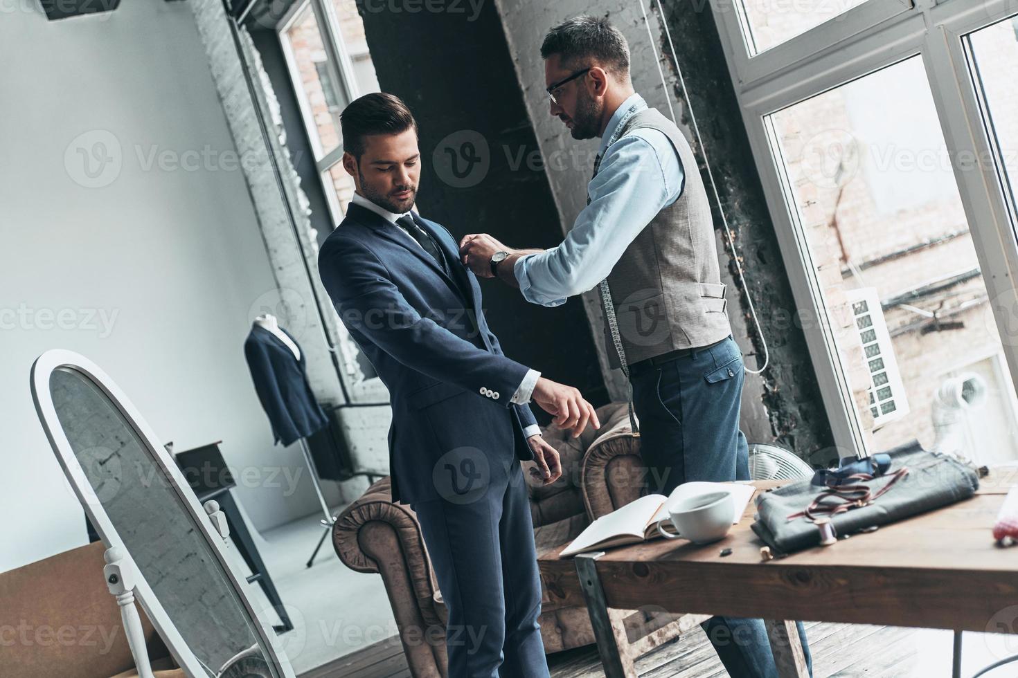 Talented tailor. Young fashionable designer helping his client to get dressed while standing in his workshop photo