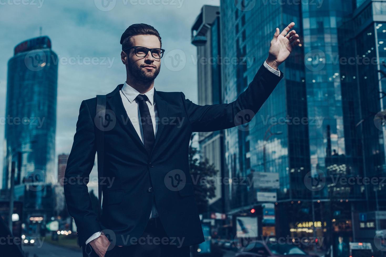 coger taxi. imagen nocturna de un joven hombre de negocios confiado con traje completo tomando un taxi mientras levanta el brazo y se para al aire libre con el paisaje urbano en el fondo foto