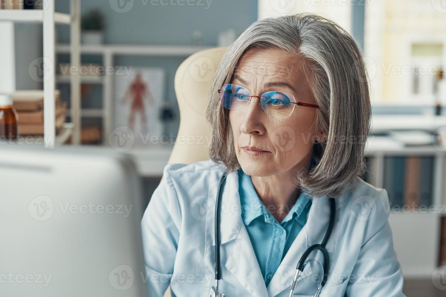 Confident mature female doctor in white lab coat working using computer while sitting in her office photo