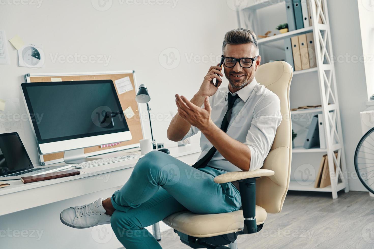 un joven apuesto con camisa y corbata hablando por teléfono inteligente y sonriendo mientras está sentado en la oficina foto