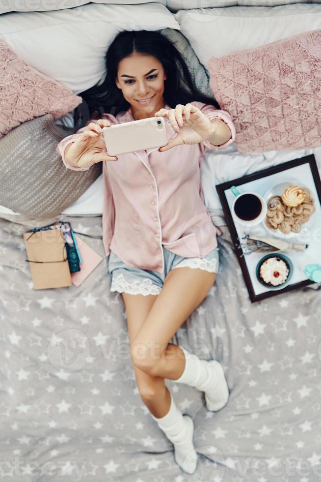 Top view of beautiful young woman in pajamas taking selfie and smiling while resting in bed at home photo