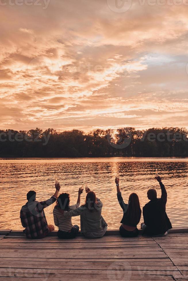 Group of young people in casual wear smiling and taking selfie while enjoying beach party near the lake photo