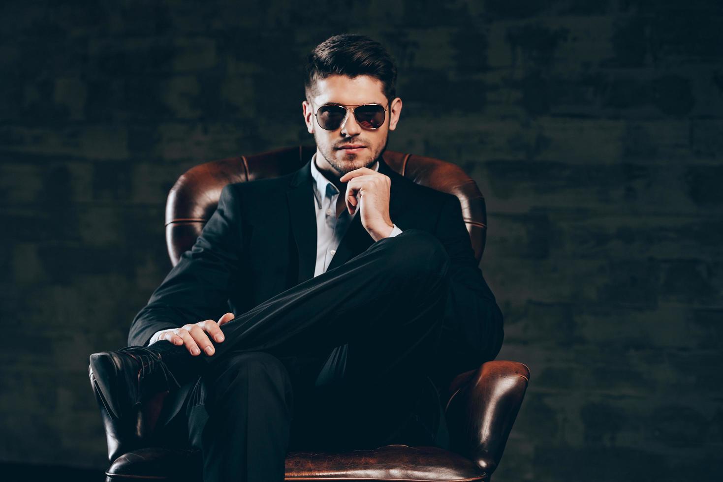 All about style. Young handsome man in suit and sunglasses holding hand on chin and looking at camera while sitting in leather chair against dark grey background photo