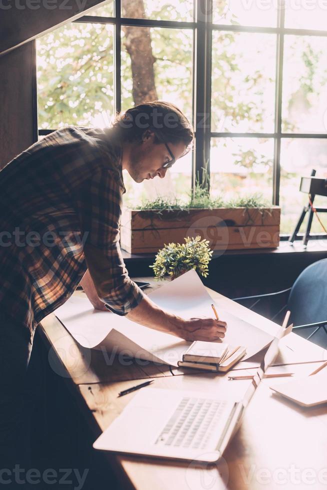 Working with passion. Confident young man in casual wear sketching on blueprint while standing near wooden desk in creative office photo