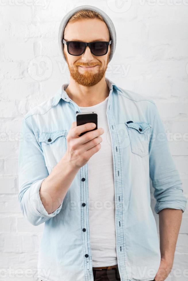 Quick text. Happy young bearded man holding mobile phone and smiling while standing against brick wall photo